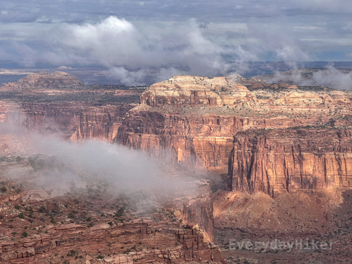 Low clouds climb over a nearby butte with a canyon and another large butte behind it. A large hill may be seen of piling rocks and sediment under the sheer cliff wall as it breaks away.