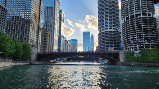 Looking straight down the Chicago river, with the famous corncob buildings on the right shore and skyscrapers on the left. in the background the sunlight illuminates some clouds and skyscrapers in the distance. the sun also reflects off the river in a single column leading from the viewer.